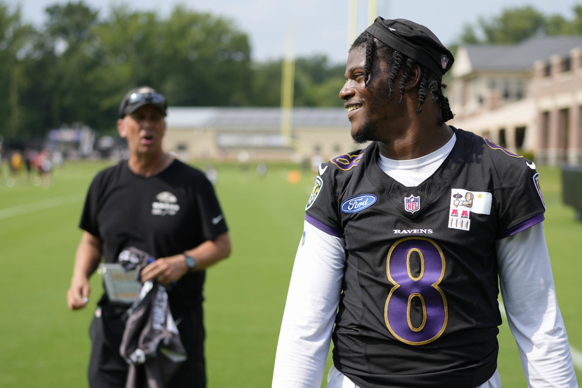 Lamar Jackson looks over his shoulder to the side and smiles with offensive coordinator Todd Monken standing in the background